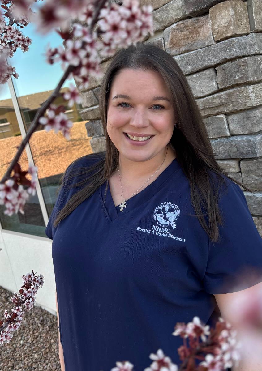 Carra Webster wearing her NNMC Nursing & Health Sciences scrubs, standing in front of a stone wall and framed by a flowering fruit tree